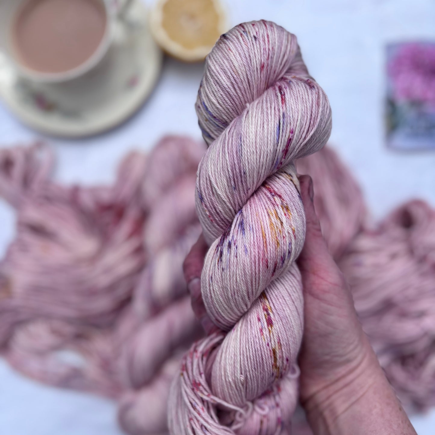 Hand holding a skein of pink yarn with a cup of tea and lemon in the background