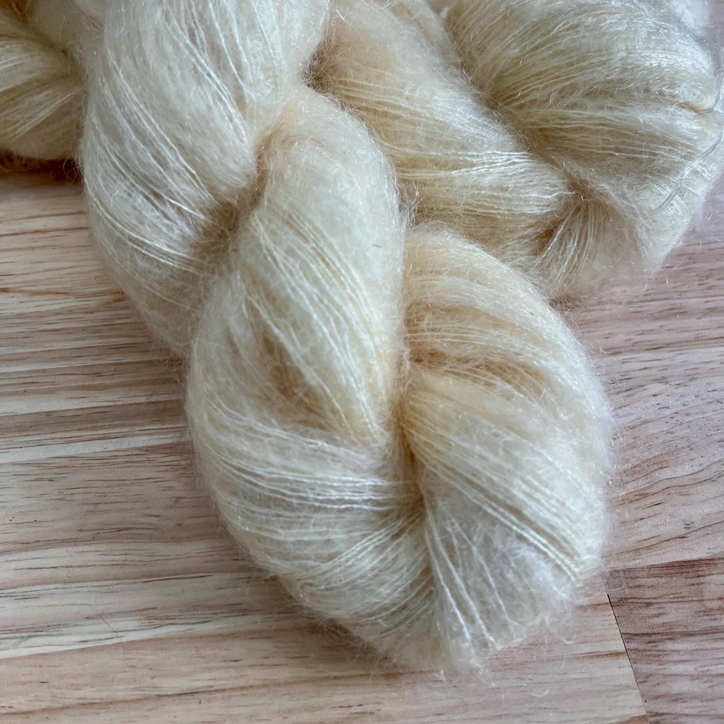 A skein of hand dyed cream colored mohair and silk yarn on a pale wood background.