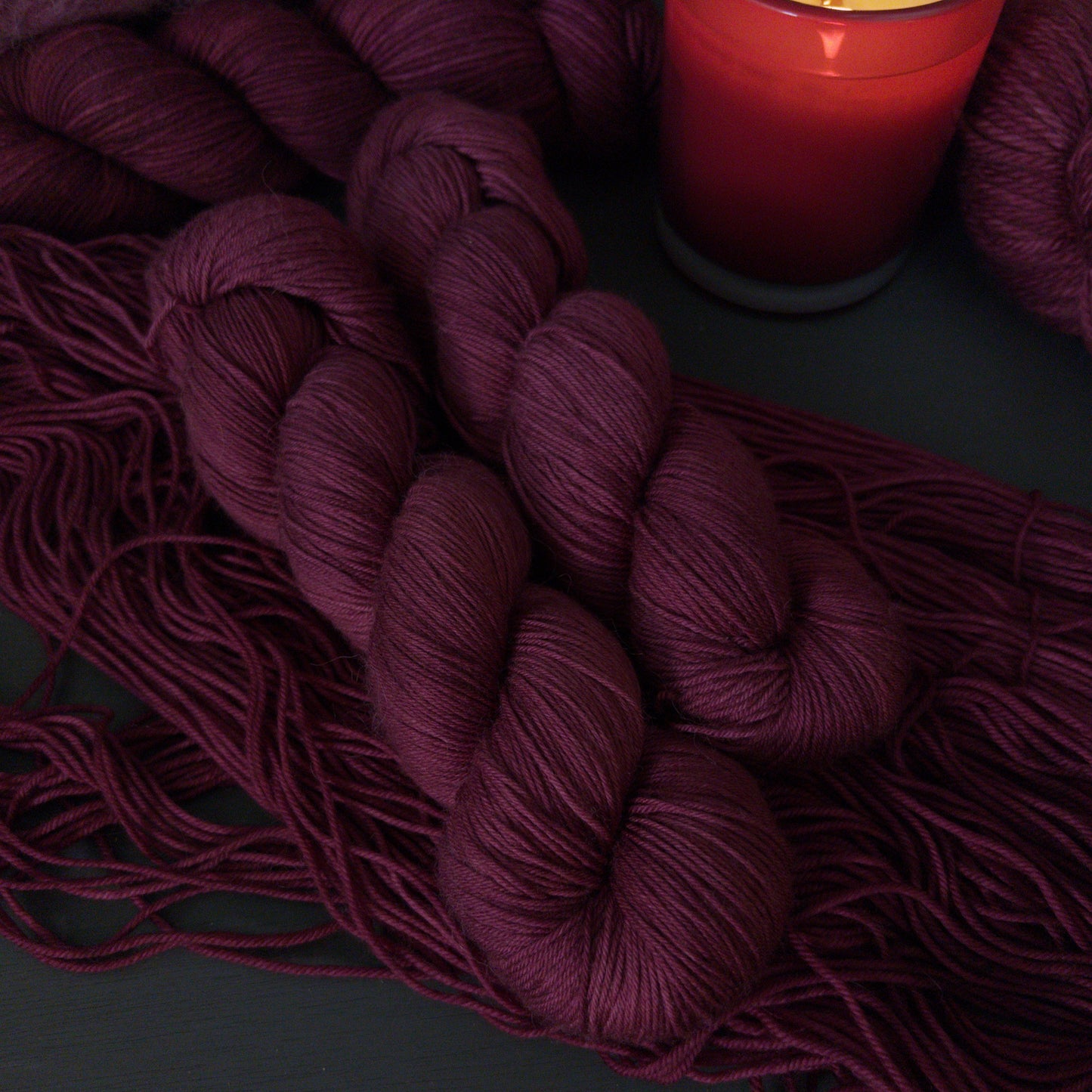 Hanks of burgundy fingering weight yarn with a lit candle on a dark surface