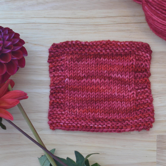 Knitted square of fabric in red and pink on a wooden surface with a flower and yarn ball.