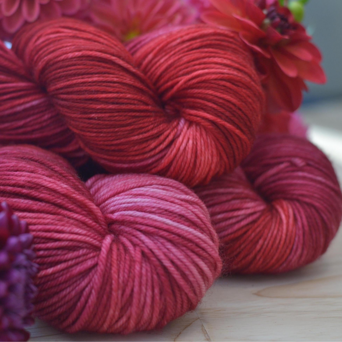 Red and pink variegated hand dyed yarn skeins on a wooden surface with flowers in the background