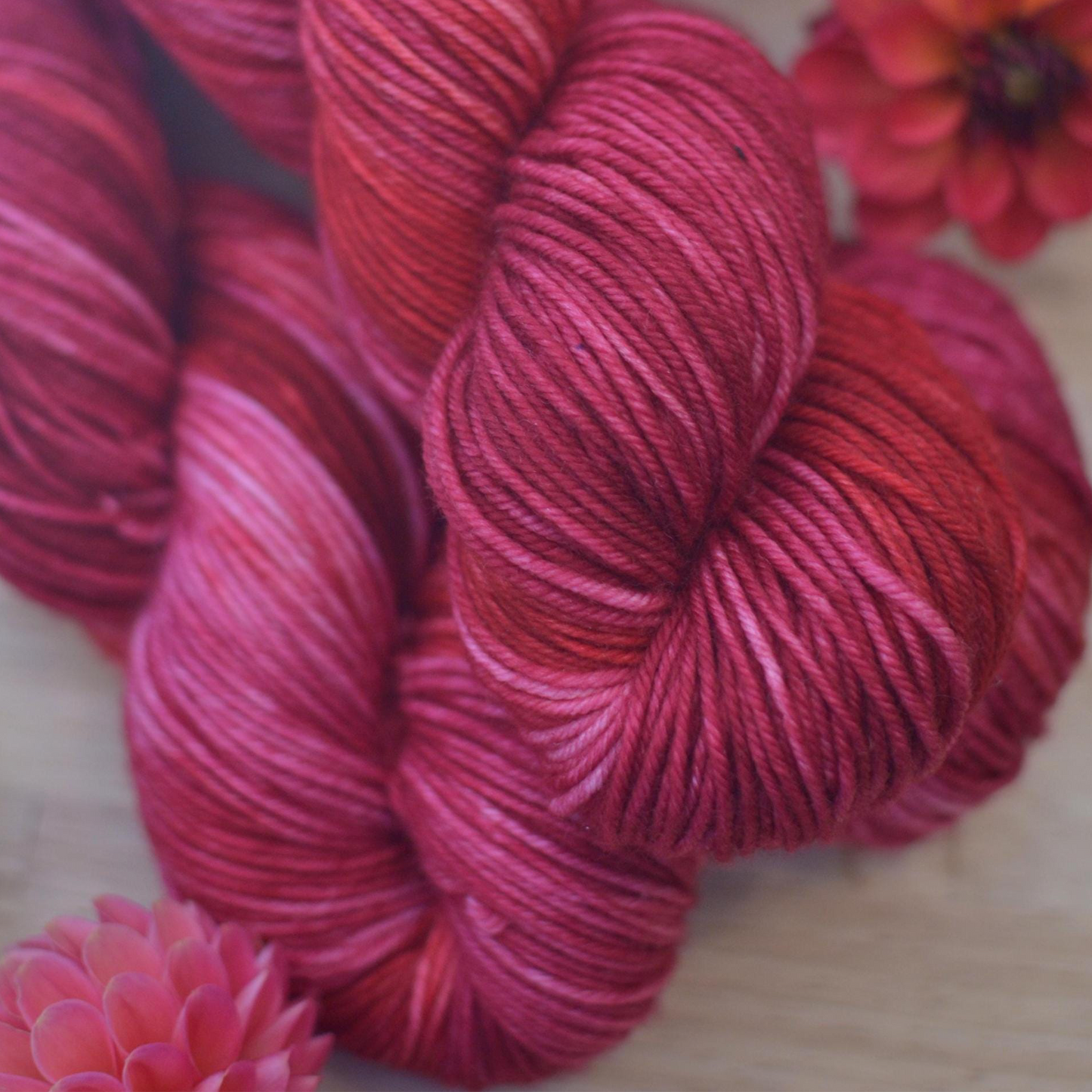 Red and pink hand dyed yarn skeins on a wooden surface with flowers.