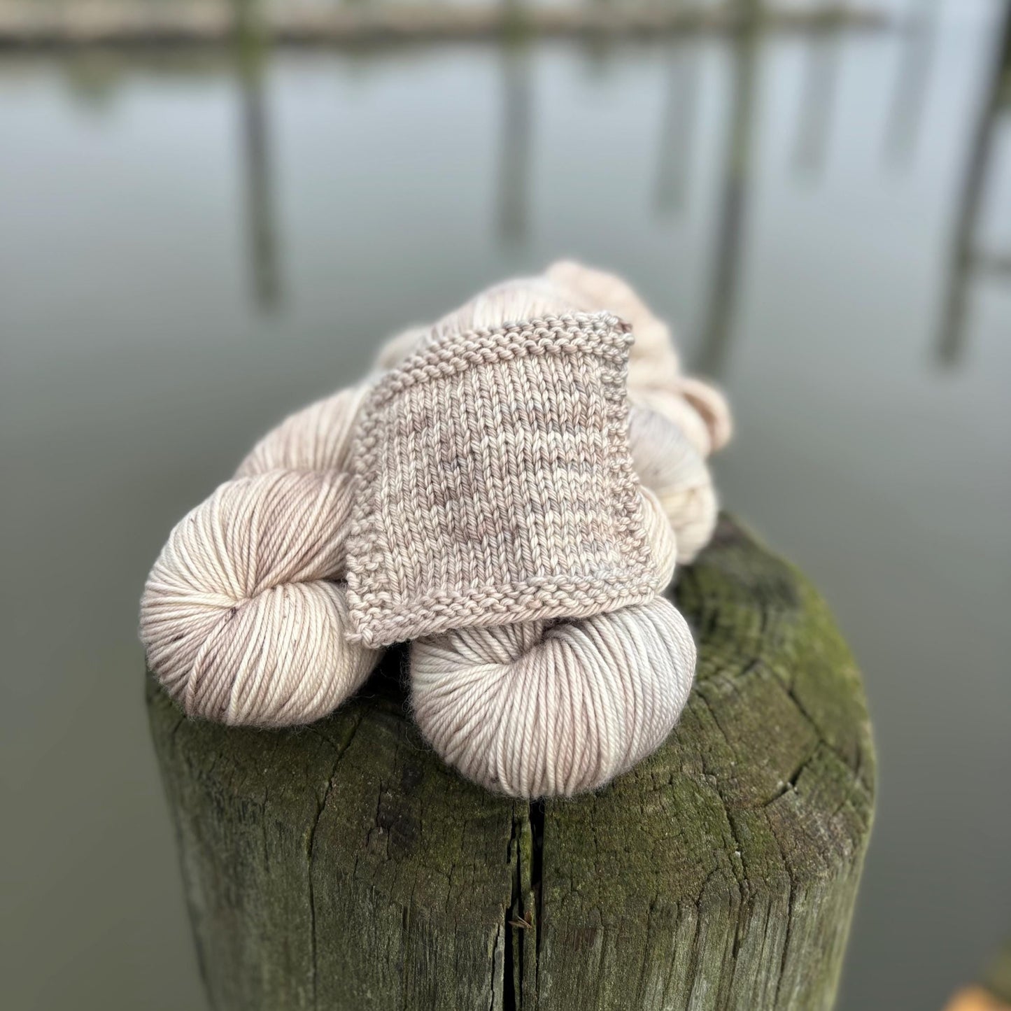 A knit swatch and skeins of hand dyed pale gray and beige yarn arranged on a pier in front of water.
