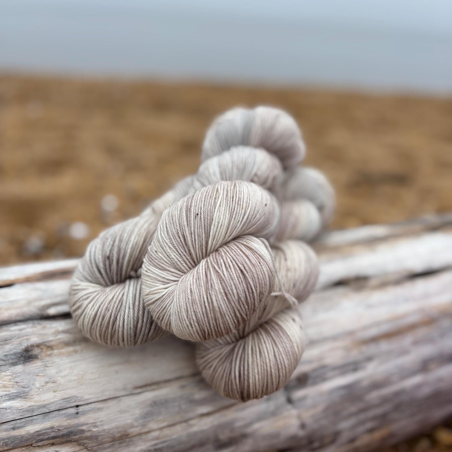Skeins of hand dyed pale gray and beige yarn arranged n a piece of driftwood.
