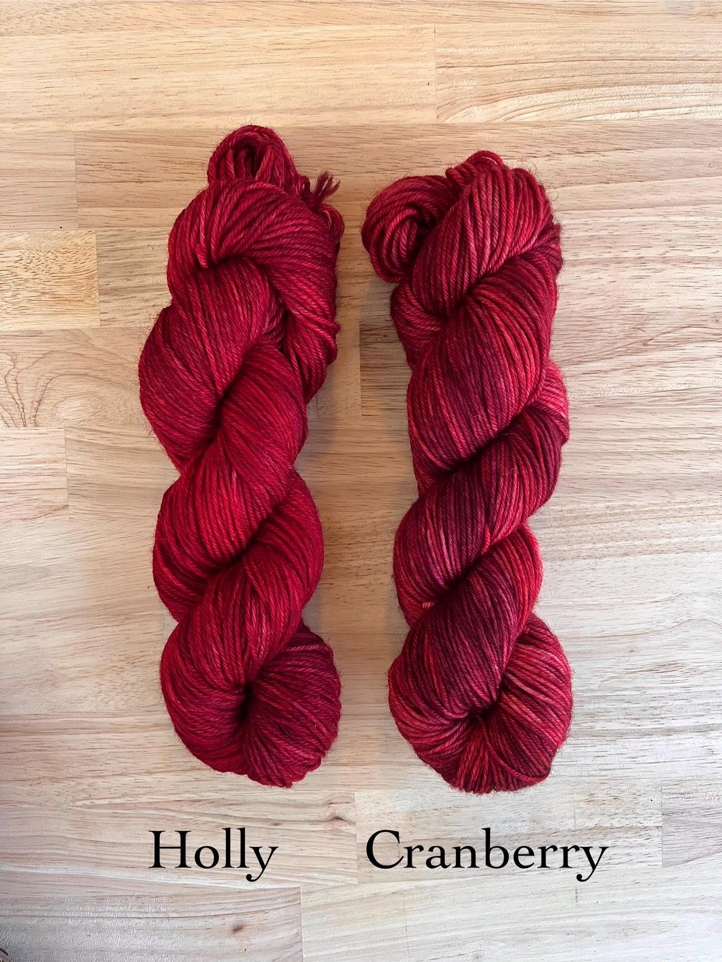 A skein of hand dyed red merino wool yarn and a skein of variegated red and burgundy hand dyed yarn arranged on a wood background.