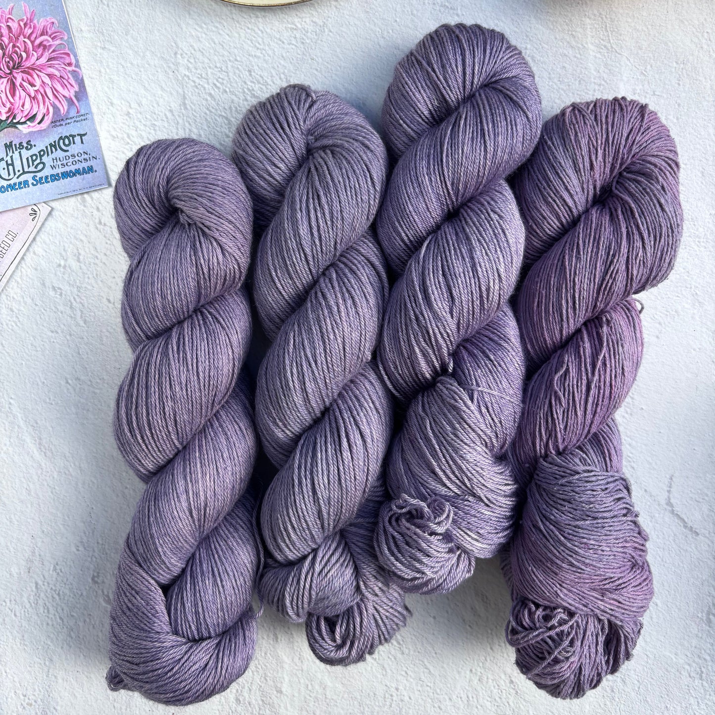 Three skeins of purple yarn on a light surface with a teacup and lemon.