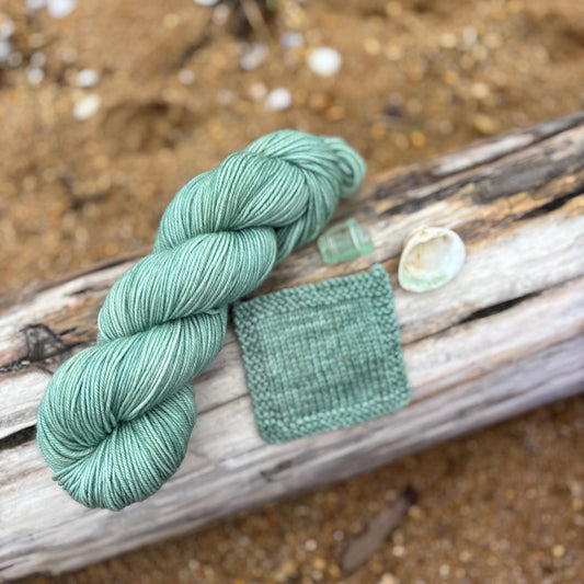 A skein and knit swatch of aqua yarn arranged on a piece of driftwood with shells placed around it.