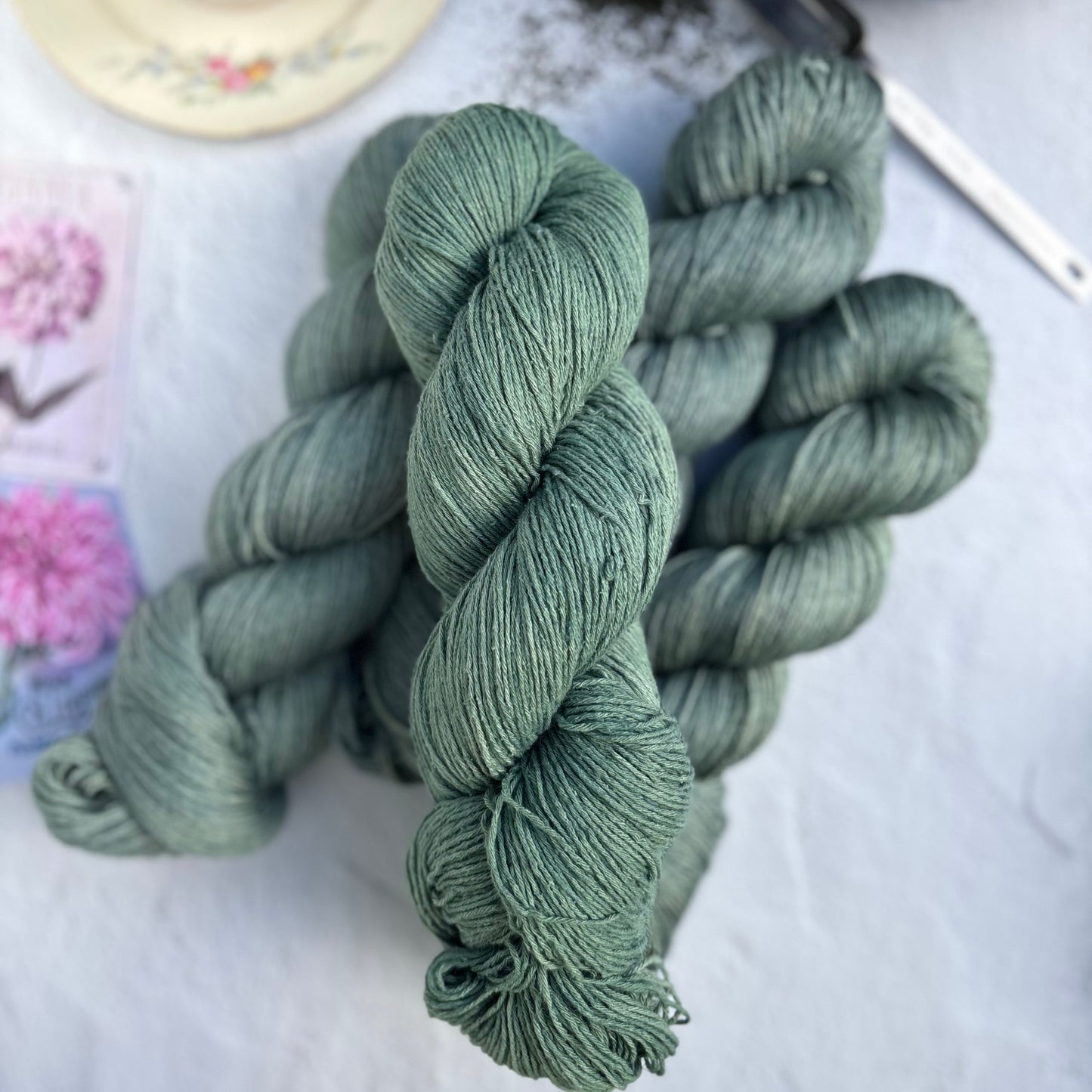 Skeins of green yarn on a light surface with a teacup and teapot in the background.