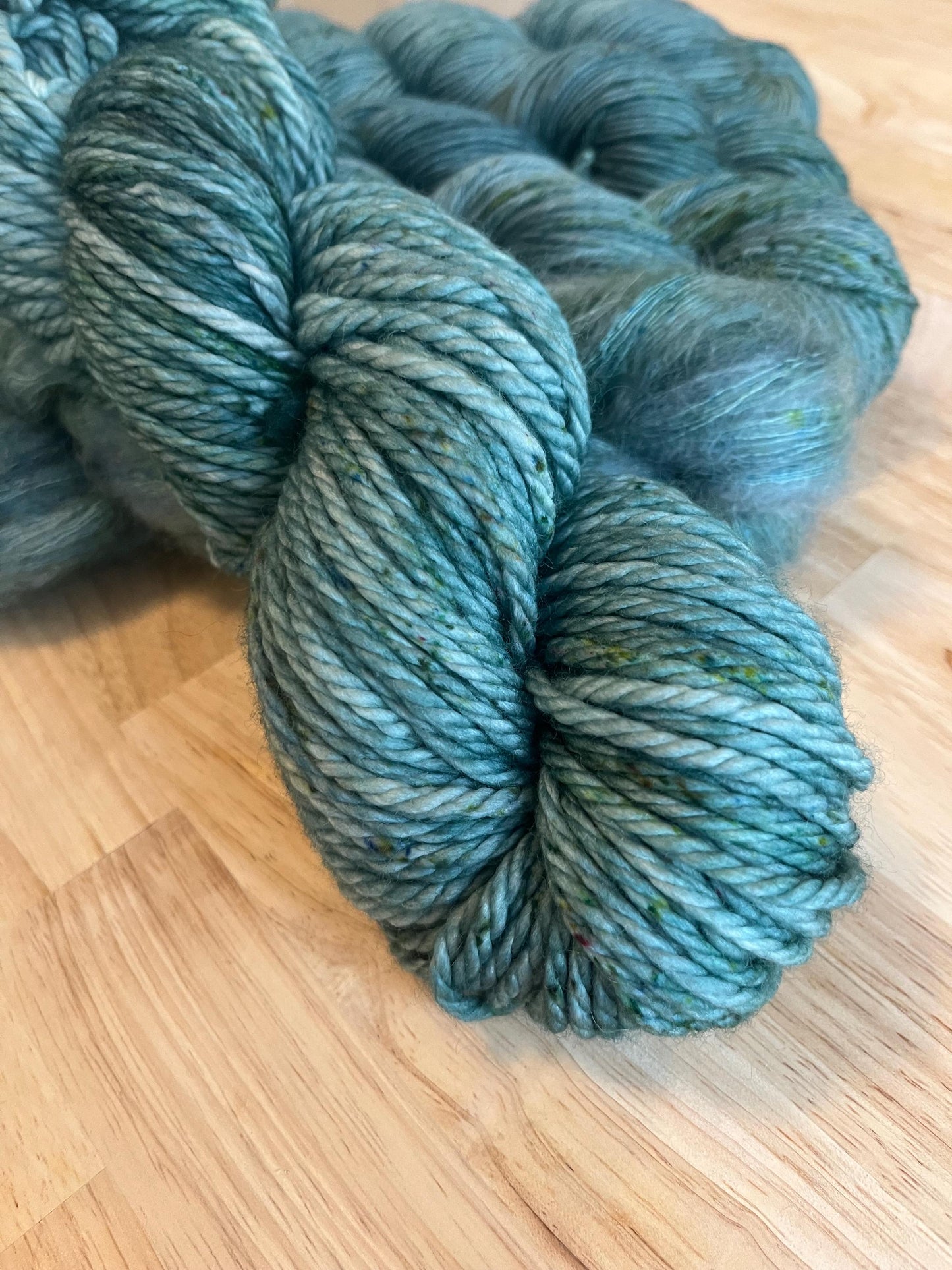 A skein of bulky weight hand dyed silvery sage green merino wool yarn with green speckles.