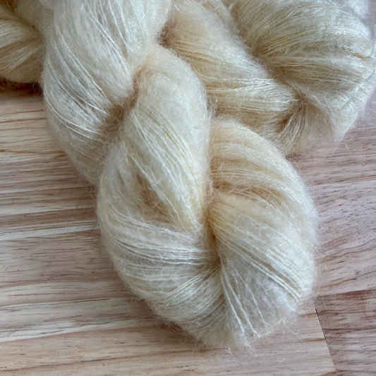 A skein of hand dyed cream colored mohair and silk yarn on a pale wood background.