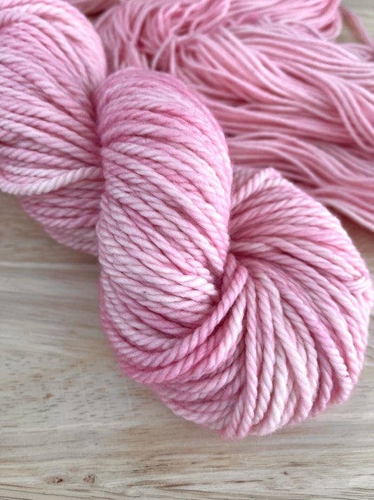 A skein of bulky weight hand dyed pale pink merino yarn arranged against a wood background.