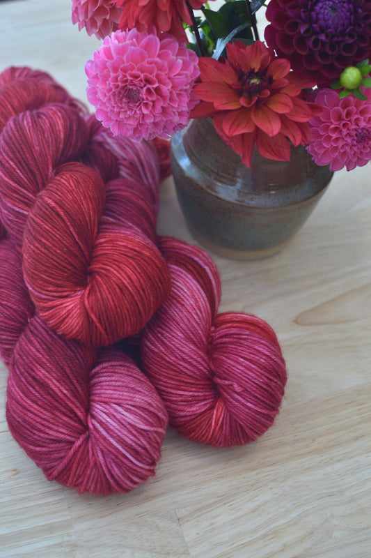 Skeins of pink and red hand dye merino wool yarn arranged with flowers.