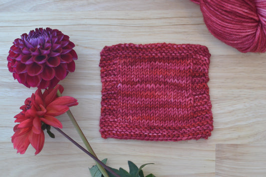 A knit swatch of hot pink and red hand dyed merino wool yarn arranged with flowers.