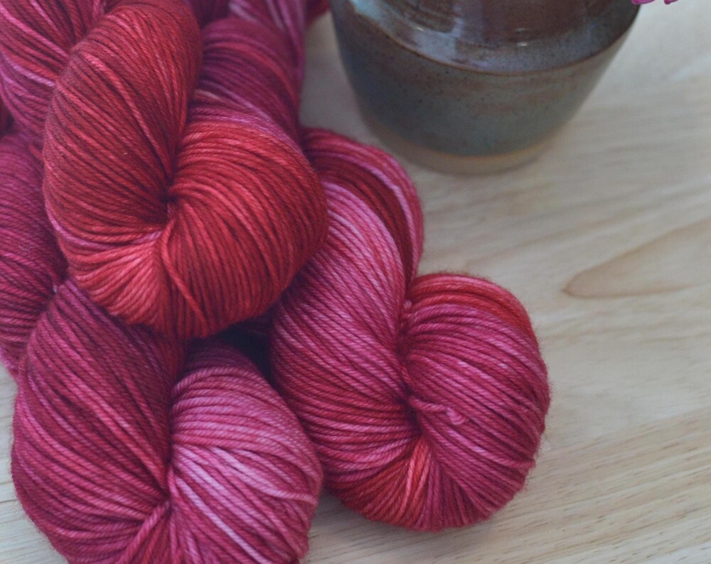 Skeins of pink and red hand dye merino wool yarn arranged with flowers.