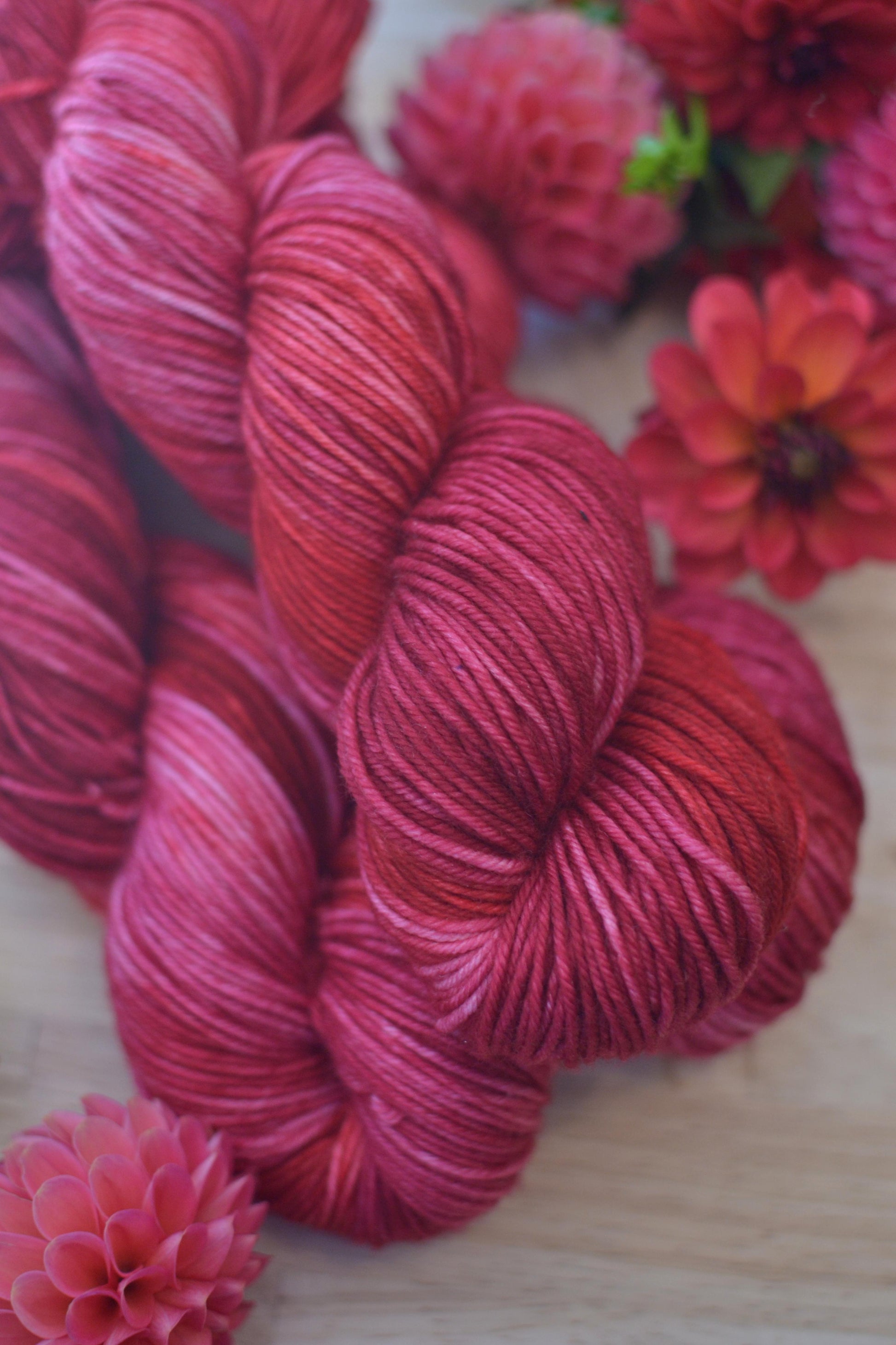 Skeins of pink and red hand dye merino wool yarn arranged with flowers.