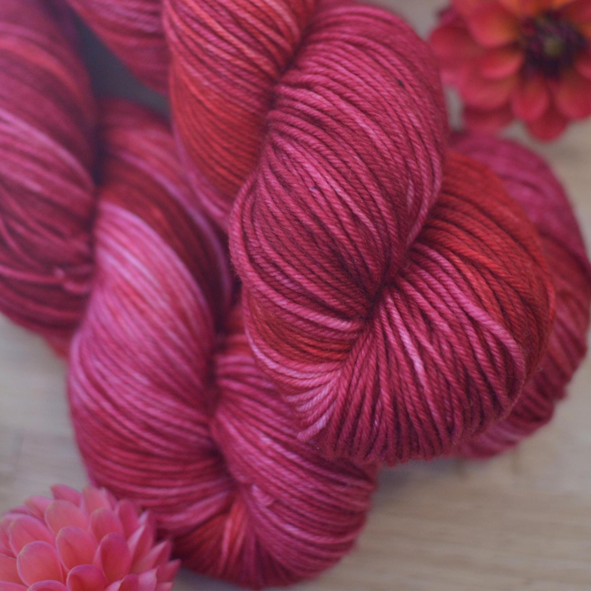 Red and pink hand dyed yarn skeins on a wooden surface with flowers.