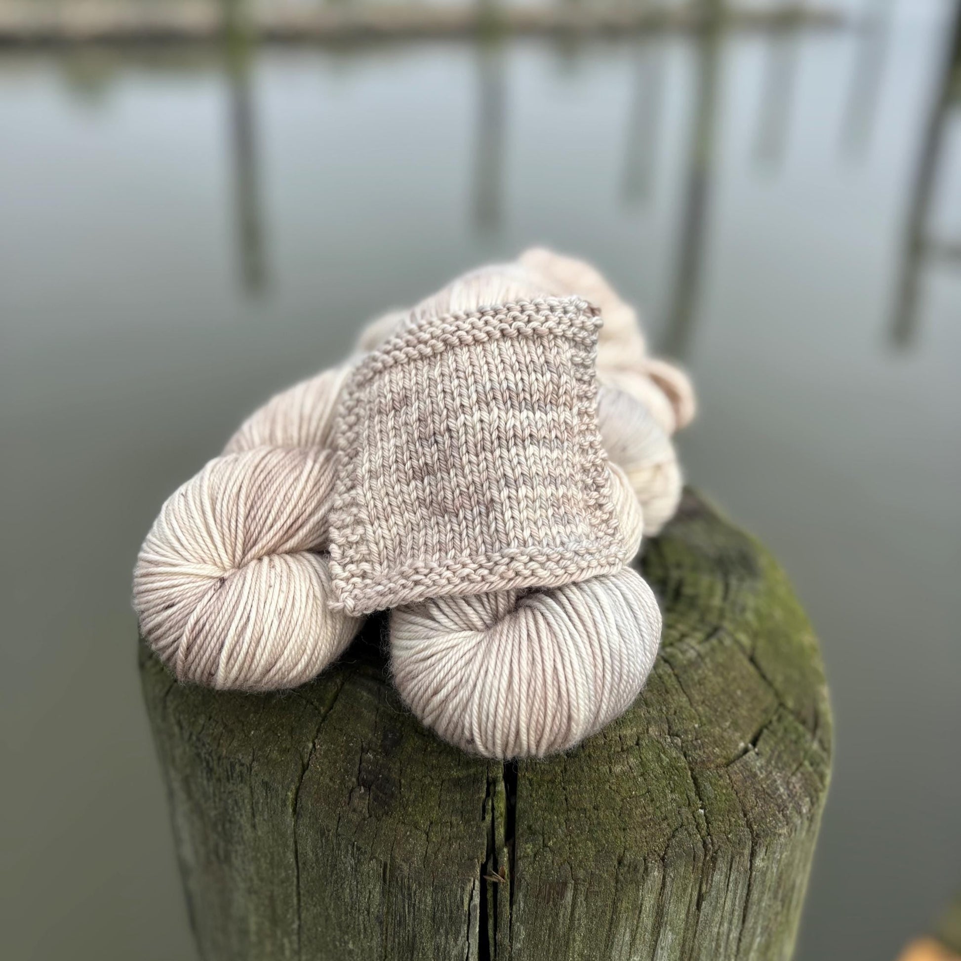 A knit swatch and skeins of hand dyed pale gray and beige yarn arranged on a pier in front of water.