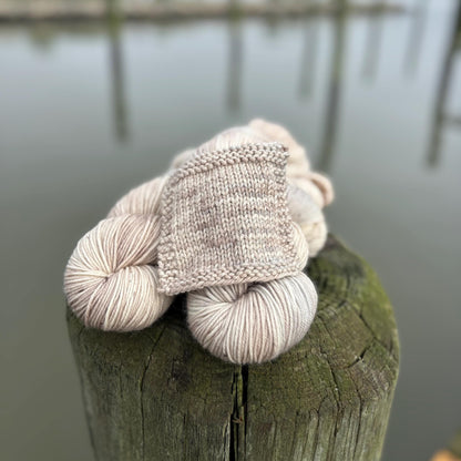 A knit swatch and skeins of hand dyed pale gray and beige yarn arranged on a pier in front of water.