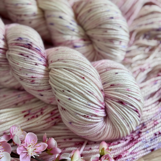 Skeins of hand dyed pale blush pink and white wool yarn with burgundy and pink speckles, displayed alongside pink flowers.