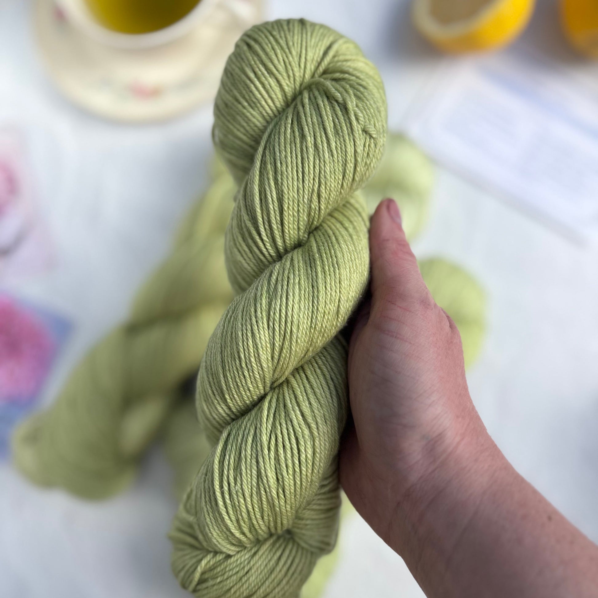 Hand holding a skein of green yarn with a lemon and other items in the background