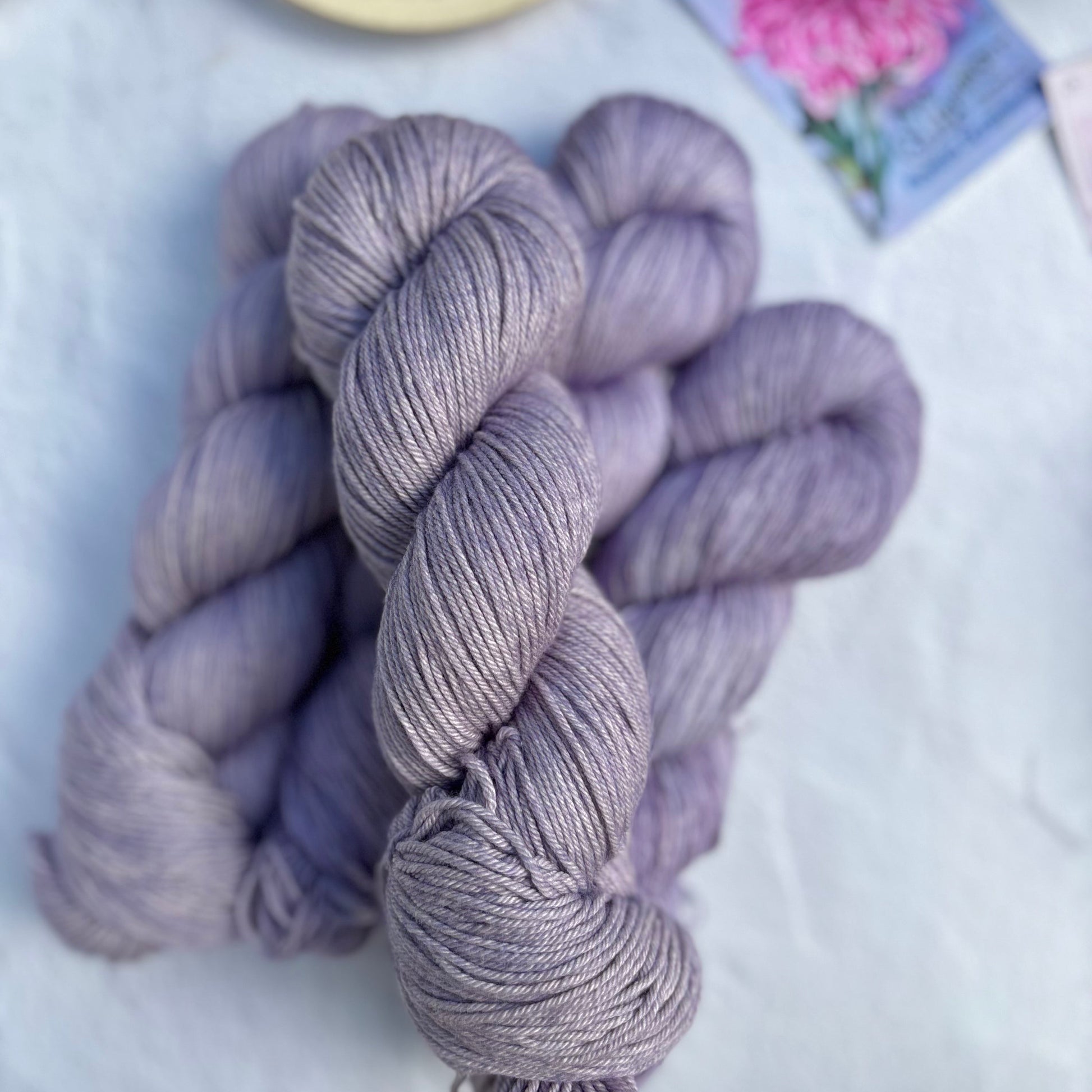 Three skeins of purple yarn on a light surface with a teacup and card in the background.