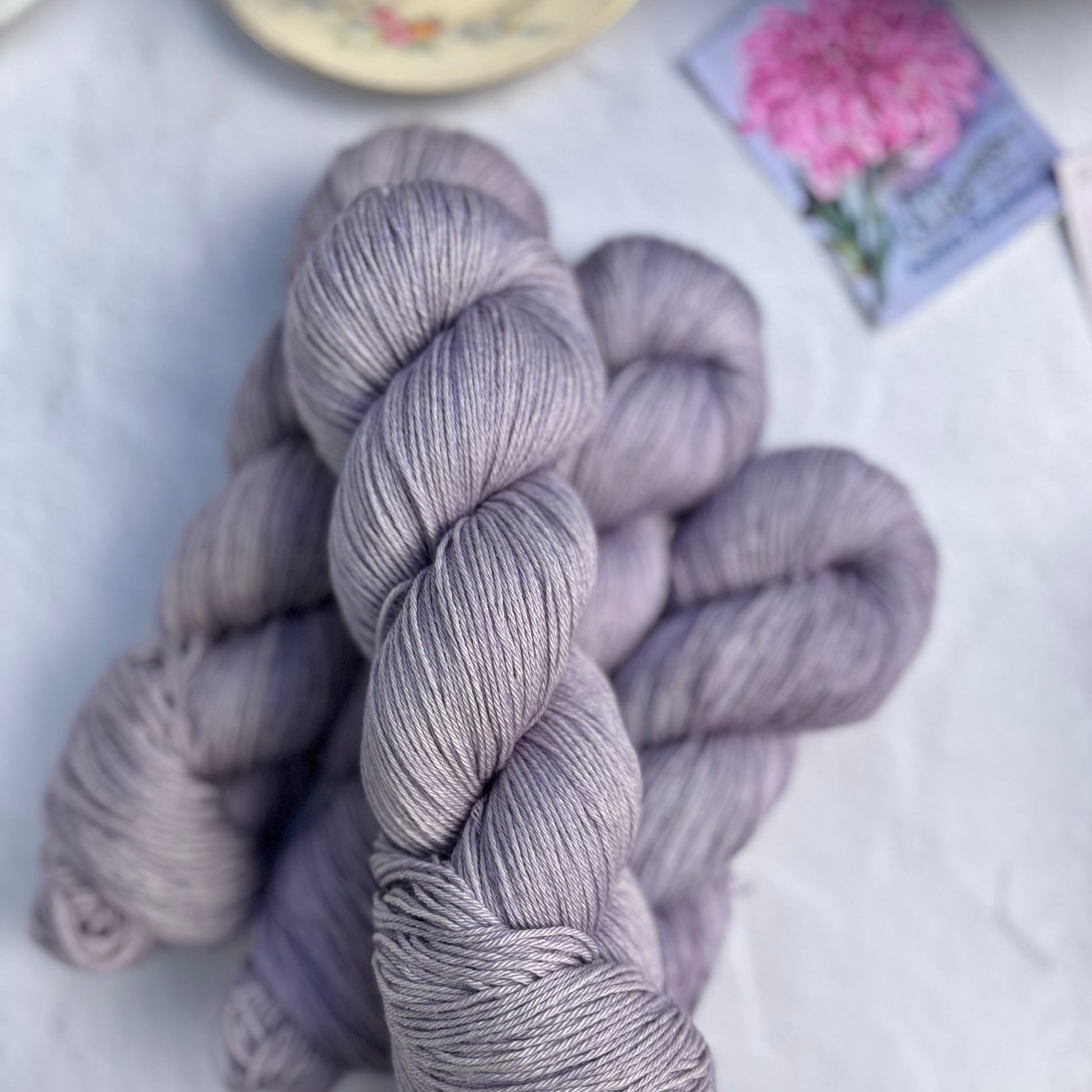 Three skeins of light purple yarn on a light surface with a teacup and pink flowers in the background.