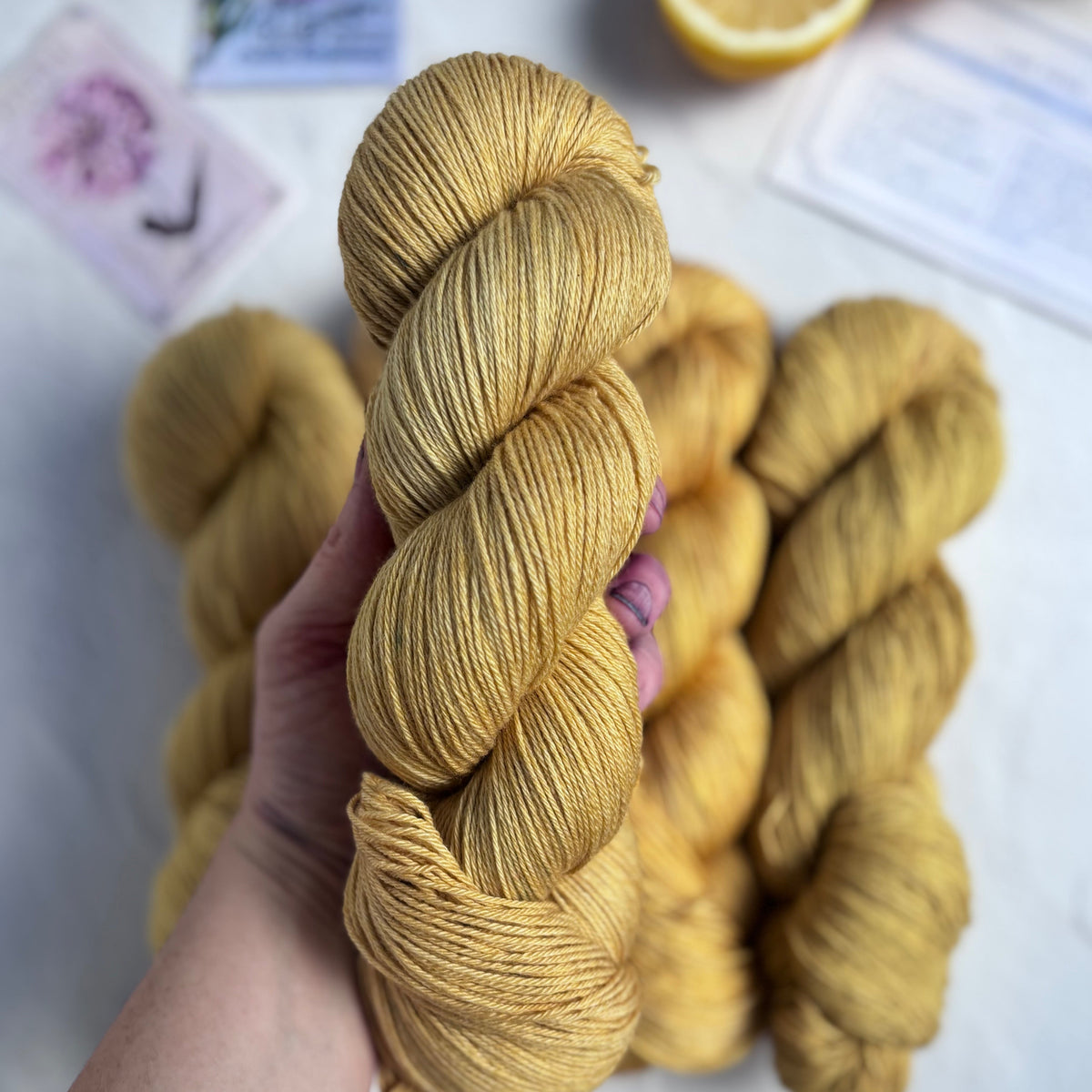 Hand holding skeins of yellow yarn with a lemon and flowers in the background
