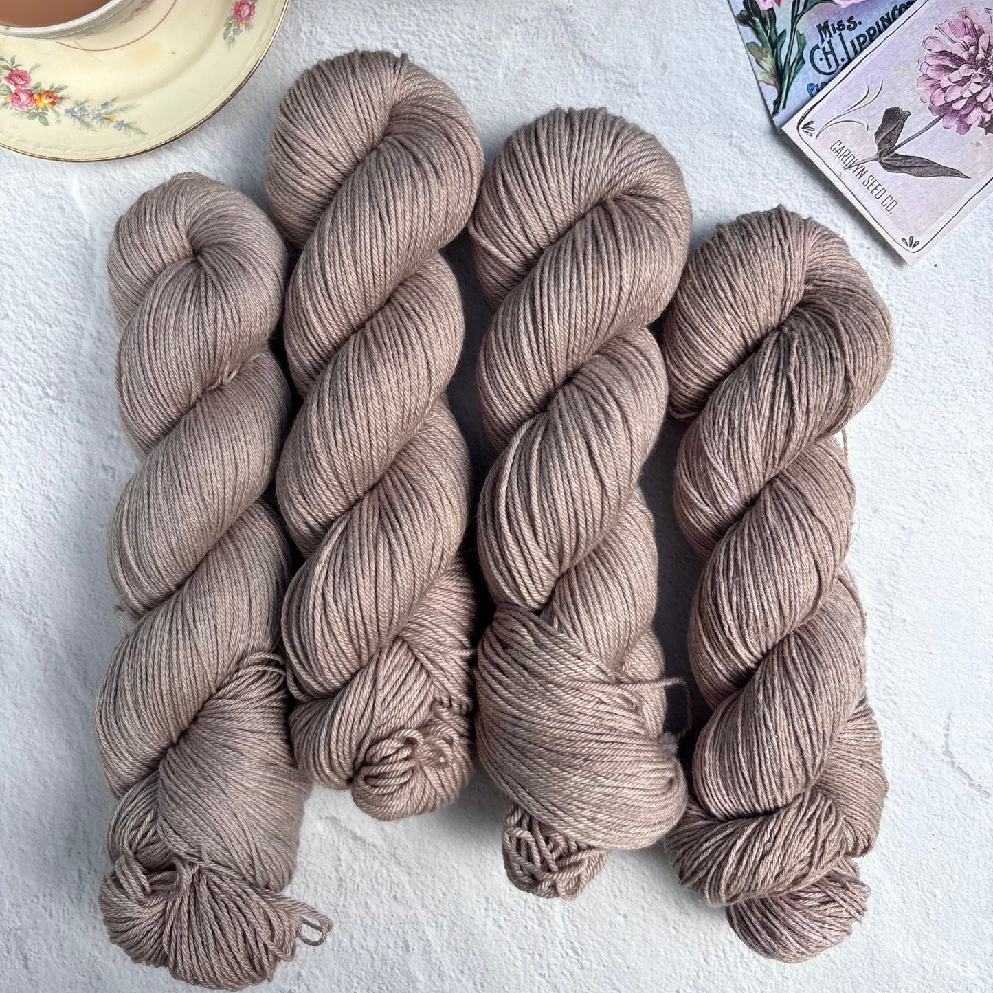 Three skeins of brown yarn on a light surface with a cup of tea and floral elements in the background.