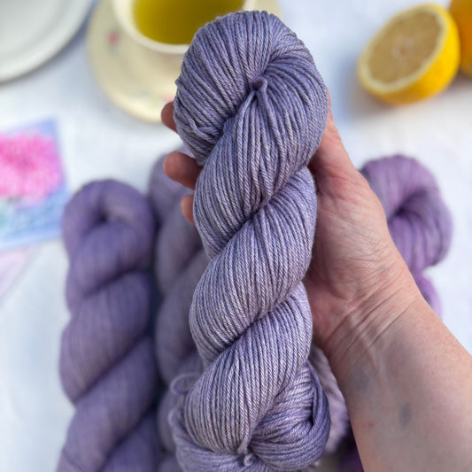 Hand holding a skein of purple yarn with more yarn and a lemon in the background.