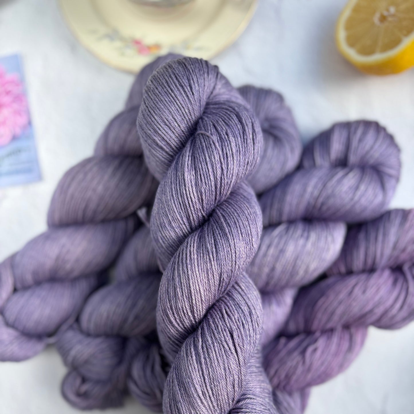 Three skeins of purple yarn on a white surface with a lemon and a colorful object in the background.