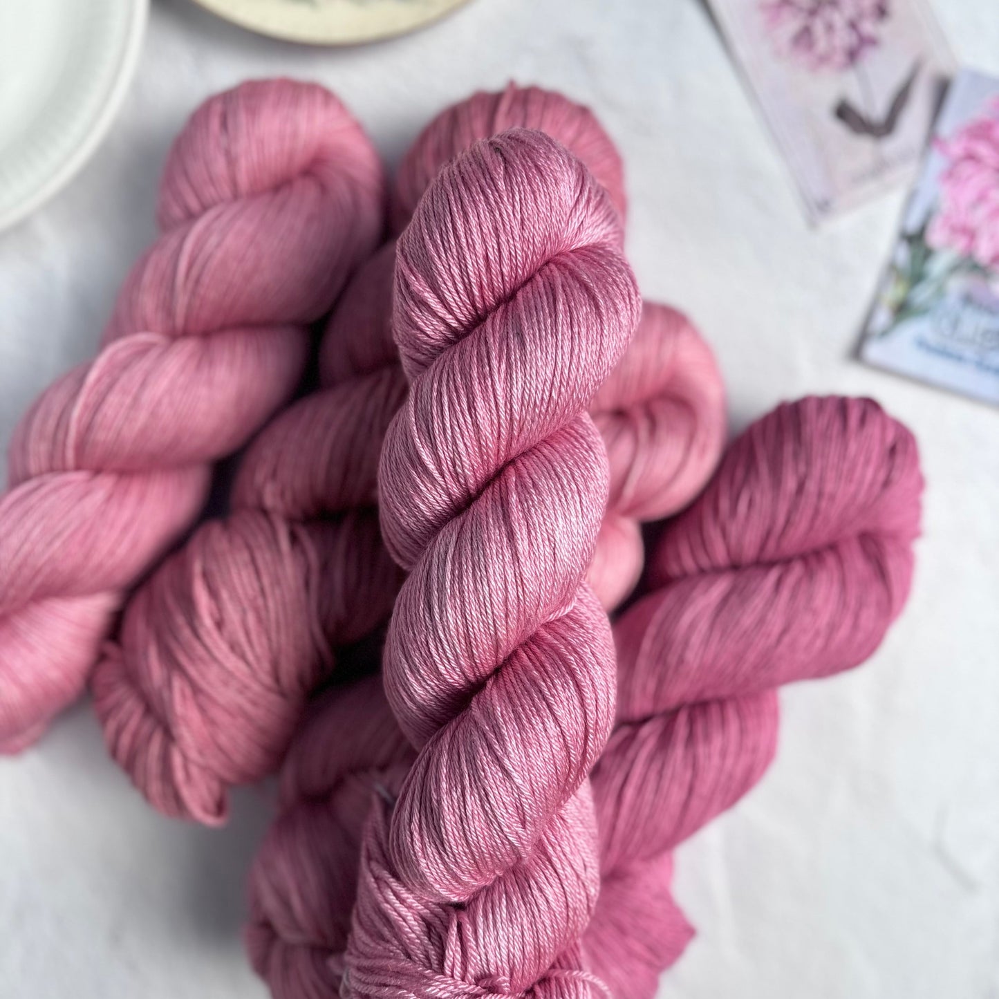 Pink yarn skeins on a light surface with a teacup and floral card in the background