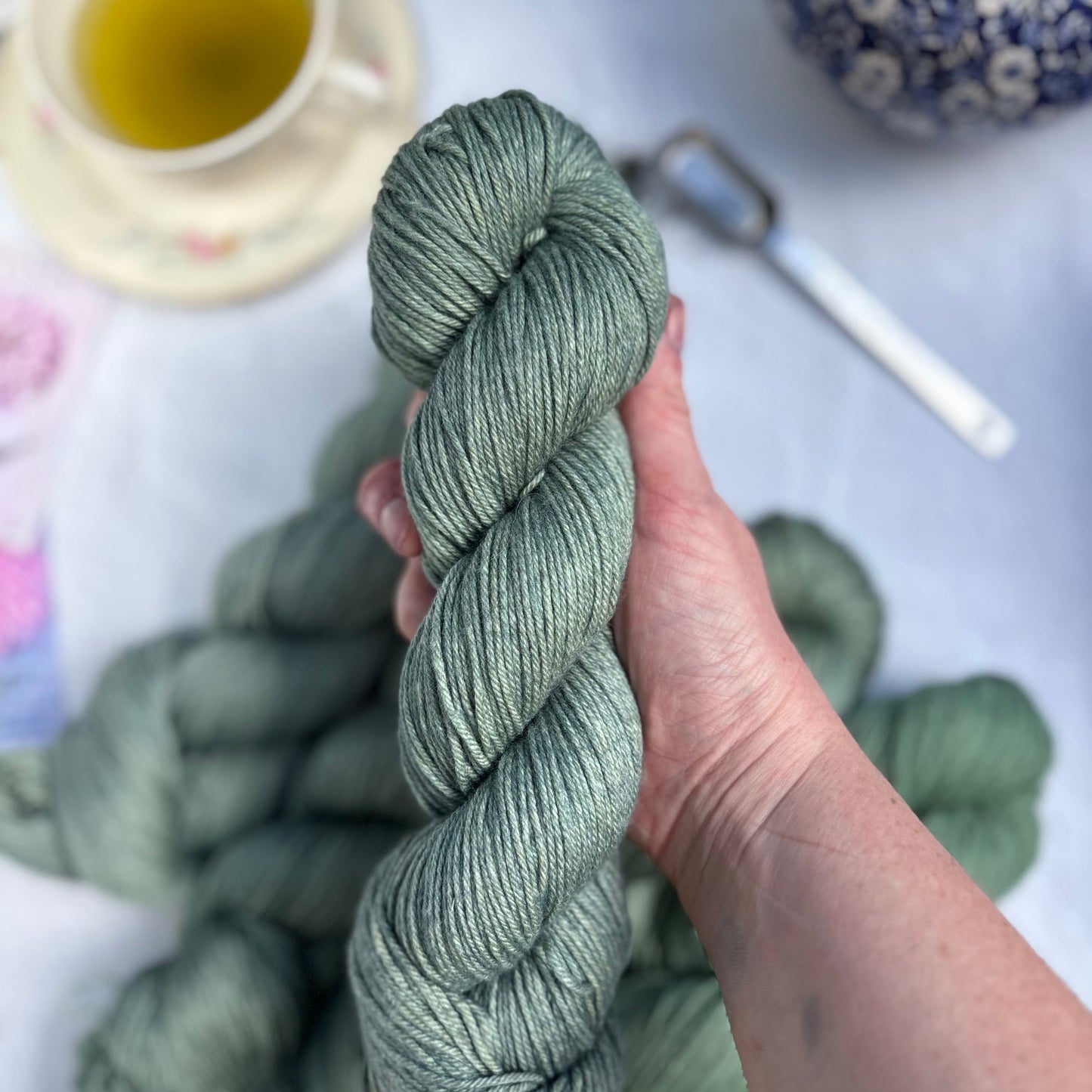 Hand holding skein of green yarn with tea set in background