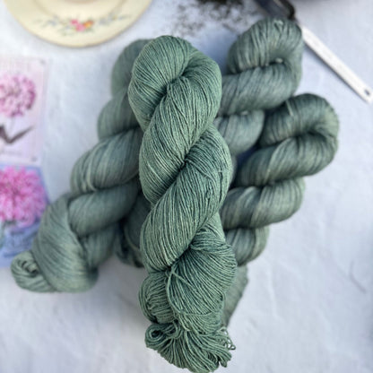 Skeins of green yarn on a light surface with a teacup and teapot in the background.
