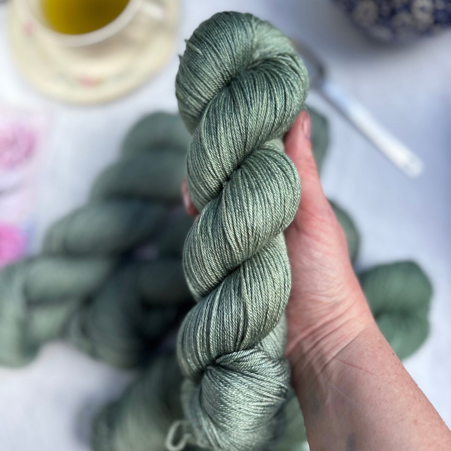 Hand holding a skein of green yarn with more yarn and a cup in the background.