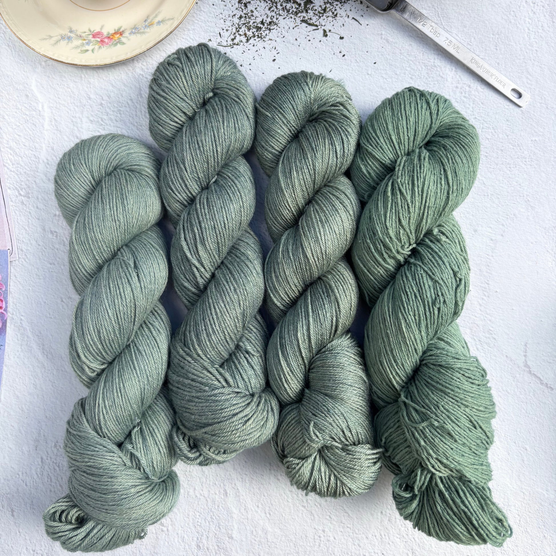 Skeins of green yarn on a white surface with a teacup and teapot in the background.