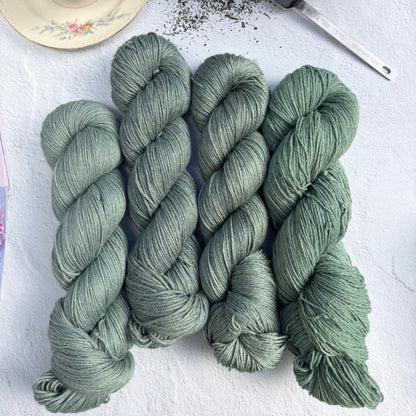Skeins of green yarn on a white surface with a teacup and teapot in the background.