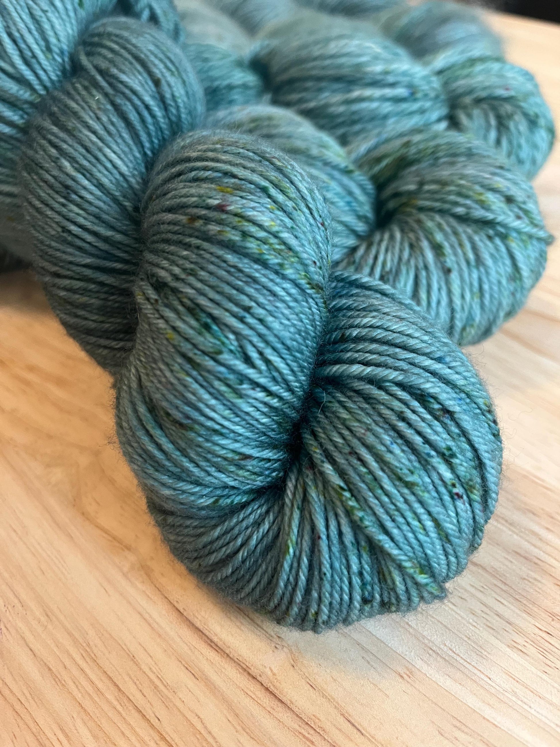 A skein of DK weight hand dyed silvery sage green merino wool yarn with green speckles.