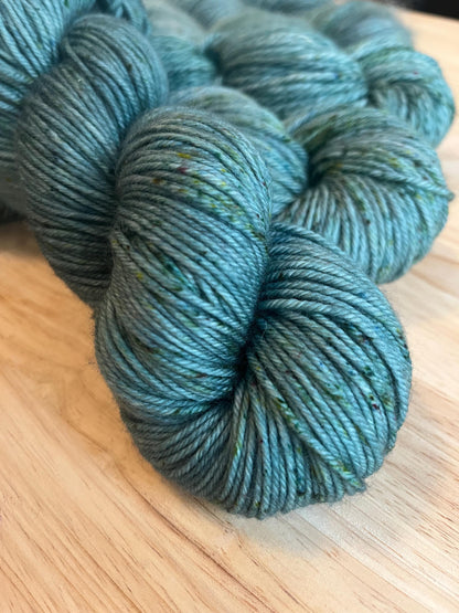 A skein of DK weight hand dyed silvery sage green merino wool yarn with green speckles.