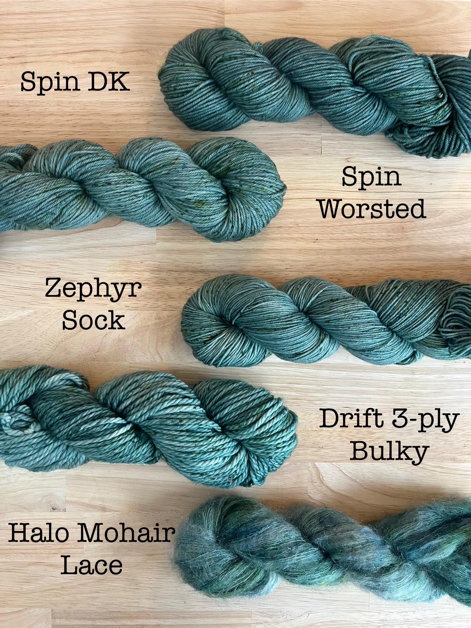 Samples of hand dyed yarn in the Spruce colorway on a variety of yarn bases including DK weight, Worsted, Sock, Bulky, and Lace. Soft gray green yarn with green speckles.