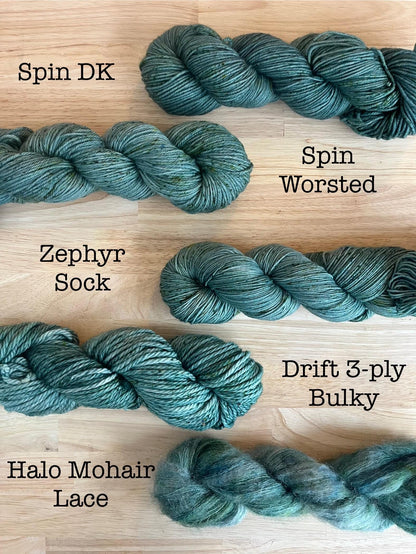 Samples of hand dyed yarn in the Spruce colorway on a variety of yarn bases including DK weight, Worsted, Sock, Bulky, and Lace. Soft gray green yarn with green speckles.