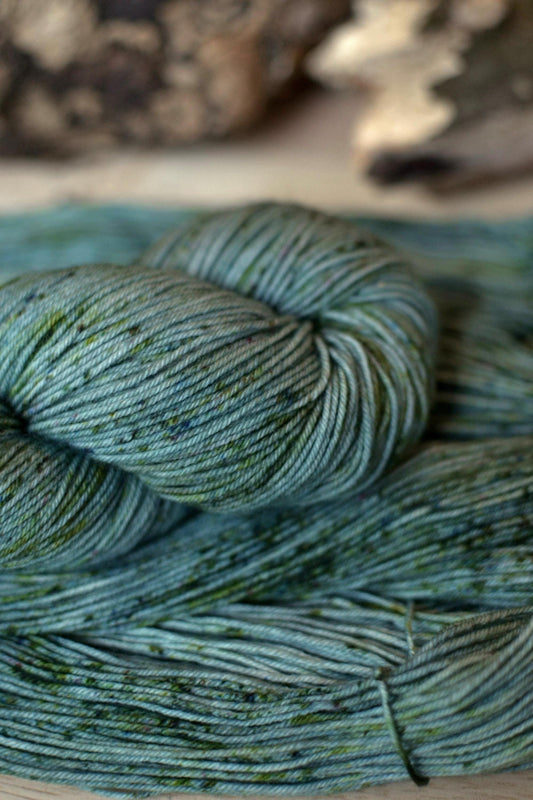 A skein of hand dyed silvery sage green merino wool yarn with green speckles.