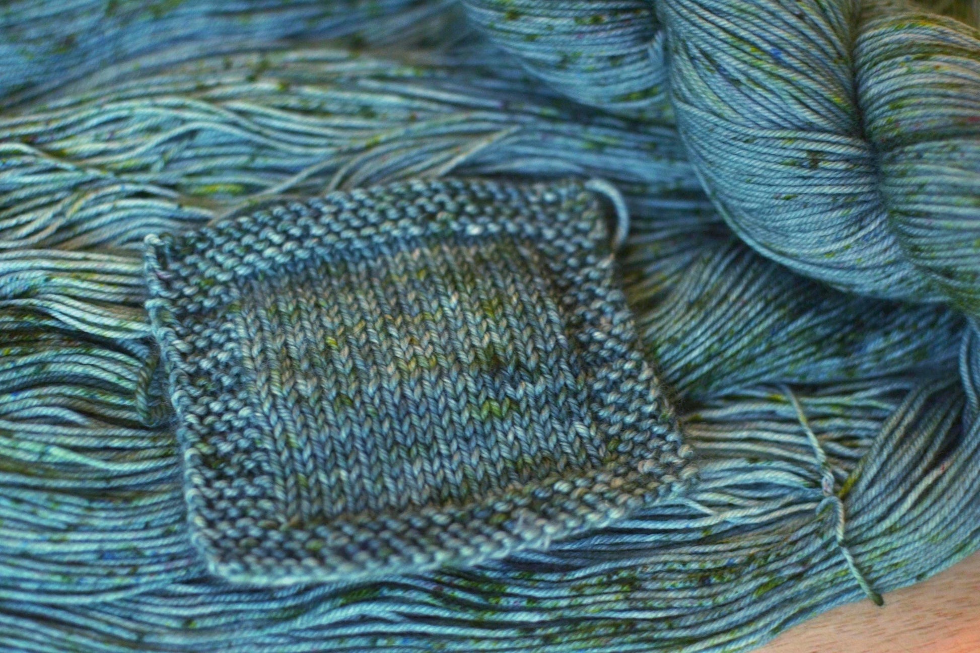 A knit swatch of hand dyed silvery sage green merino wool yarn with green speckles.