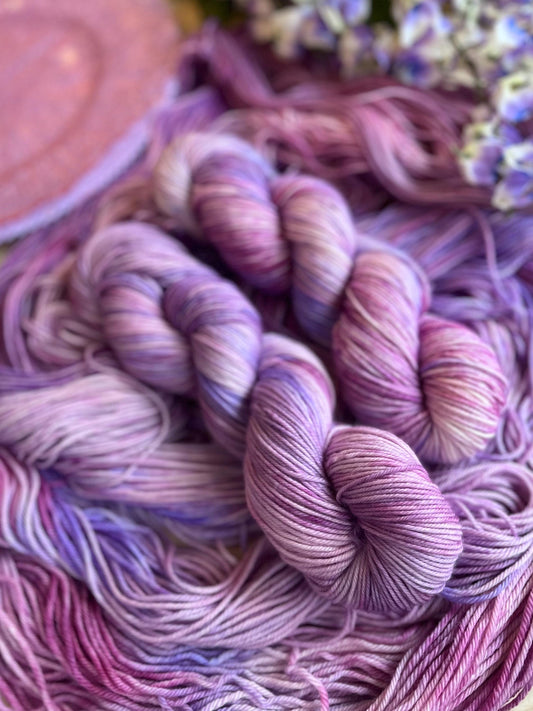 Skeins of hand dyed pink and purple merino wool yarn arranged with flowers.