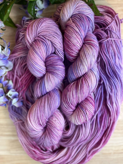 Skeins of hand dyed pink and purple merino wool yarn arranged with flowers.