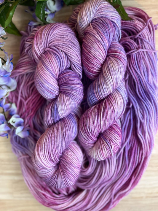 Skeins of hand dyed pink and purple merino wool yarn arranged with flowers.