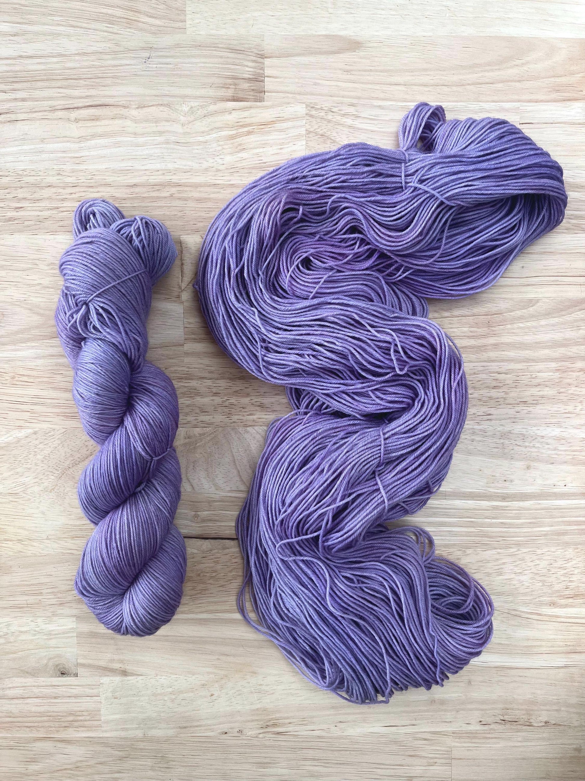 Skeins of sock weight light purple hand dyed merino yarn against a wooden background.