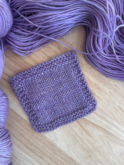 A knit swatch of hand dyed light purple merino yarn against a wooden background with skeins of wool yarn arranged around it.