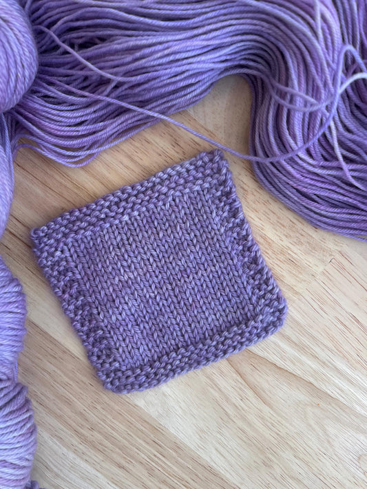 A knit swatch of hand dyed light purple merino yarn against a wooden background with skeins of wool yarn arranged around it.